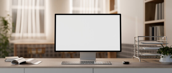 A white-screen computer mockup and office supplies on a table in a cozy contemporary office.