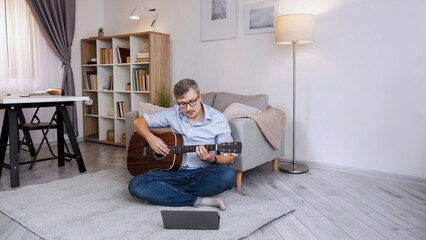 Musician student. Online lesson. Home education. Smart concentrated man practicing acoustic guitar chords sitting at laptop video meeting on floor in living room copy space.