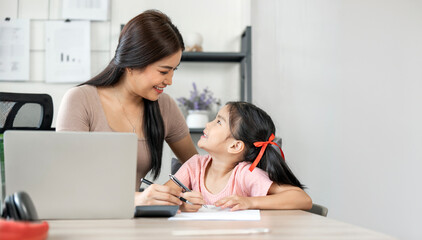 Beautiful business mom helping her little daughter doing home work while working from. home.