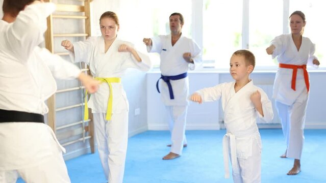 Boy with his family in kimonos and colored belts practicing karate with punches during group martial arts class in gym, accompanied by trainer