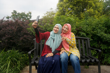 Asian woman with her friends sitting on bench and taking selfie together using mobile phone at park