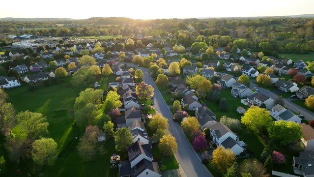 Springtime sunset in American housing development. Aerial view of a suburban neighborhood at sunset, featuring rows of houses surrounded by lush, colorful trees.