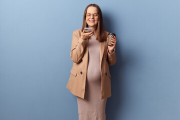 Delighted joyful smiling young adult pregnant woman wearing dress and jacket holding mobile phone...
