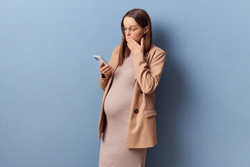 Surprised young adult pregnant woman wearing dress and jacket posing isolated over blue background...