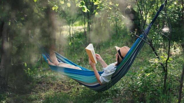 A young Caucasian woman in a hat reading a book while lying in a hammock in a spring garden