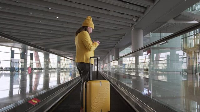 Busy modern woman with suitcase at airport on travelator looks at phone app online for boarding pass and flight information.