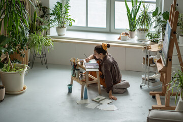 Creative woman artist sits on floor, painting in cozy art studio with houseplants. Drawing process...