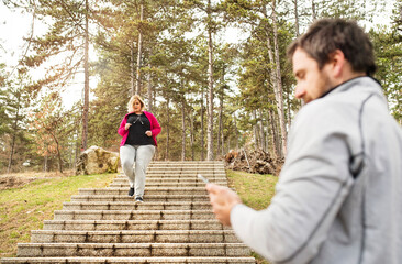Overweight woman running down the stairs, personal trainer checking her performance. Exercising outdoors for people with obesity, support from friend, fitness coach.