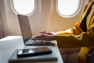 Young Asian business woman in yellow suit uses laptop sitting near window on airplane to do online...