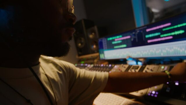 Close-up tilting of hand and face of busy black male audio engineer in headphones sitting in control room during recording session, moving up feeder slider and turning equalizer knob on mixing desk