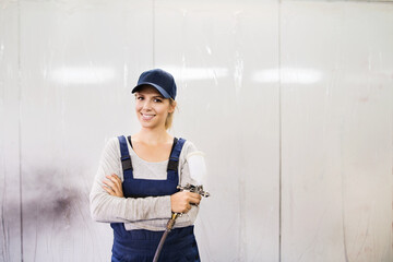 Female car painter, paingting vehicles in auto body shop. Young woman holding spray gun, looking at camera, smiling.