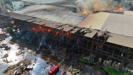A bird's-eye perspective reveals a sprawling industrial edifice ravaged by fire. Billowing smoke...