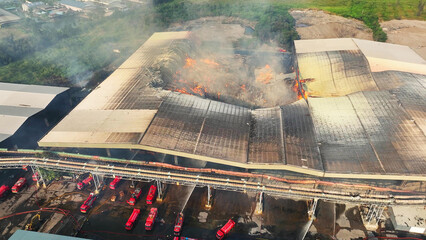 From above, an industrial inferno rages, fire trucks battle fiercely to contain the blaze, smoke...