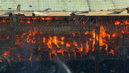 Devastation reigns as flames devour an industrial complex, the collapsed roof a stark foreground to...
