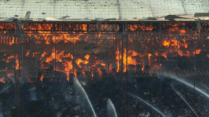 In the foreground, a collapsed roof bears witness to a raging industrial fire, flames leaping...