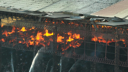 A raging inferno consumes an industrial site, its destructive flames dancing amidst collapsed roofs. Smoke engulfs the sky, a testament to the devastation below.
