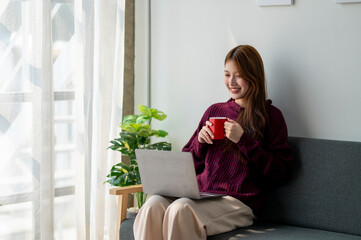 A woman is sitting on a couch with a laptop and a red coffee mug. She is smiling and she is...