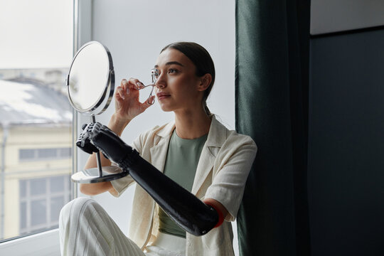 Young woman with bionic arm looking at herself in mirror using eyelash curler