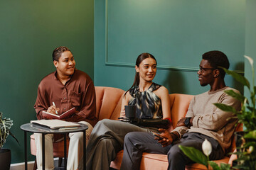 Smiling diverse colleagues discussing over laptop sitting on sofa in office
