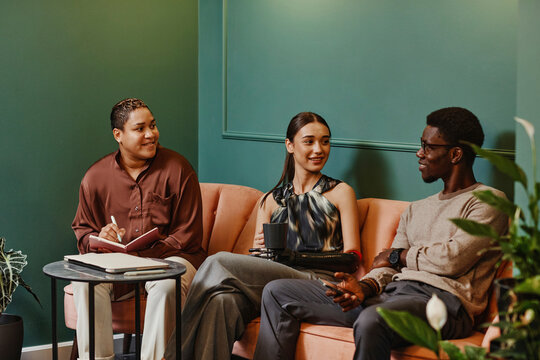 Smiling diverse colleagues discussing over laptop sitting on sofa in office