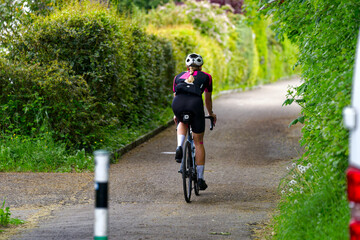 Female cyclist with blond hair pigtail on racing bicycle surrounded by bushes on a rural road at...