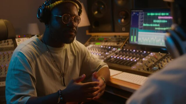 Medium over-shoulder of cheerful African American male sound engineer with headphones sitting in recording studio, talking to record label producer and discussing ways to improve new track