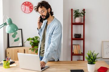 Confident young bearded creative man talking on phone while using laptop at studio office.