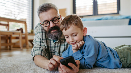 Little boy watching cartoon movie on smartphone with father, lying on floor in kids room. Dad...