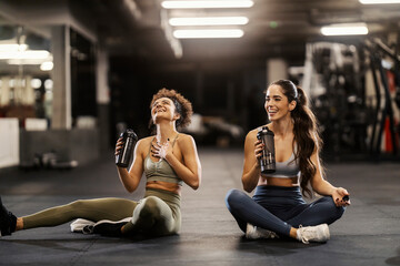 Two happy sporty friends sitting on a gym floor and drinking water while taking a break.