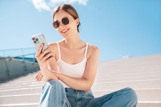 Young beautiful smiling hipster woman in trendy summer casual clothes. Carefree woman posing in the street. Positive model holds mobile phone, looks at cellphone screen, uses smartphone apps