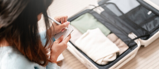Fototapeta premium luggage, clothes, suitcase, baggage, tourism, journey, person, room, list, packing. a woman is writing on a piece of paper and is holding a pen. The suitcase is open and has clothes inside.