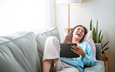 enjoyment, lying, cute, rest, living, cozy, pretty, relaxing, couch, comfortable. A woman is sitting on a couch with a tablet in her lap. She is smiling and laughing while using the tablet.
