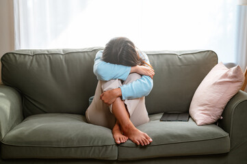 anxiety, children, cry, depressed, despair, frustration, grief, mental, pain, regret. A woman is sitting on a couch, crying and holding her stomach. She is wearing a blue shirt and white pants.