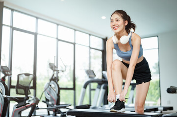 Fototapeta premium fit, fitness, sport, exercise, gym, muscle, muscular, physical, vitality, wellness. A woman is in a gym, wearing headphones and a blue tank top. She is bending over to tie her shoe.