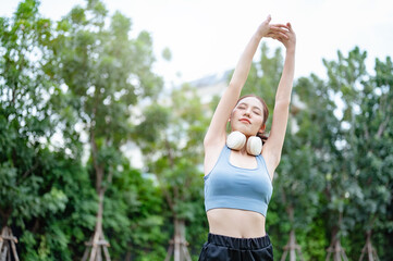 meditation, fit, relaxation, pilates, stretch, runner, vitality, wellbeing, wellness, posture. A woman is stretching her arms in the air while wearing headphones. She is in a park surrounded by trees.