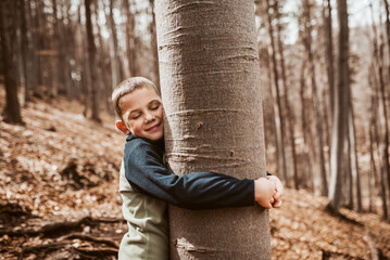 Cute boy on walk in forest, hugging a tree, enjoying the peaceful moment, his eyes closed. Child...
