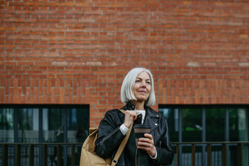 Portrait of stylish mature woman with gray hair on city street. Older woman in leather jacket with soft smile.