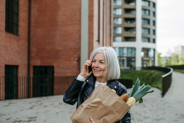 Fototapeta premium Mature woman making phone call, going home from supermaket with gorceries. Beautiful older woman with gray hair standing on city street.