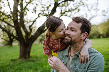 Daughter giving father small flower, kissing him on cheek. Dad and girl spending together time in spring nature. Father's day concept.