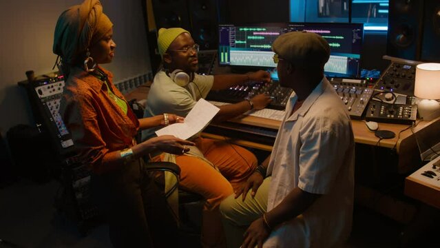 Medium full of young stylish Black female singer in turban standing in control room, talking to male producer and audio engineer, while preparing to record vocal tracks for new album