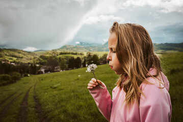 Girl blowing a dandelion in spring nature, rain clouds on sky. Wearing rain jacket, side view. Spring or autumn weather.