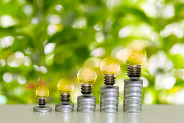 The image depicts a light bulb placed on top of a stack of coins, set against a nature background....