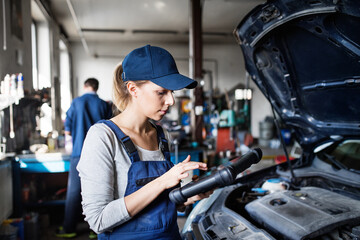 Female auto mechanic using diagnostic tool, scanner, running diagnostic on car. Beautiful woman working in a garage, wearing blue coveralls.