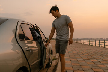 Asian man closing car door, parking a car next to the river at sunset time after long drive, happy...