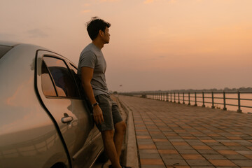 Asian man standing relaxing and leaning the car look at nature at sunset time, looking out the river, happy travel concept.