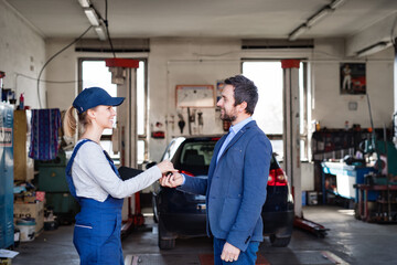 Female auto mechanic talking with customer, handing over keys of repaired car. Beautiful woman working in a garage, wearing blue coveralls.