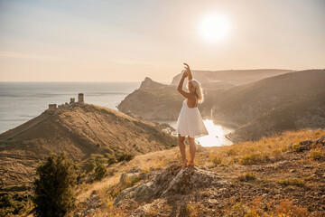 A woman stands on a hill overlooking a body of water. She is wearing a white dress and she is enjoying the view.