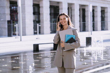 Smiling businesswoman on a phone call, holding documents, confidently walking through a sunny city plaza with fountains in the background