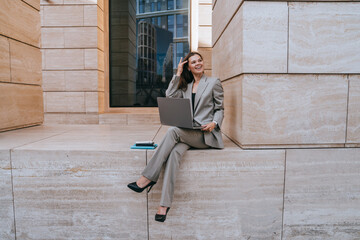 Relaxed and smiling businesswoman working on her laptop outdoors, comfortably seated on a stone step in a modern urban setting