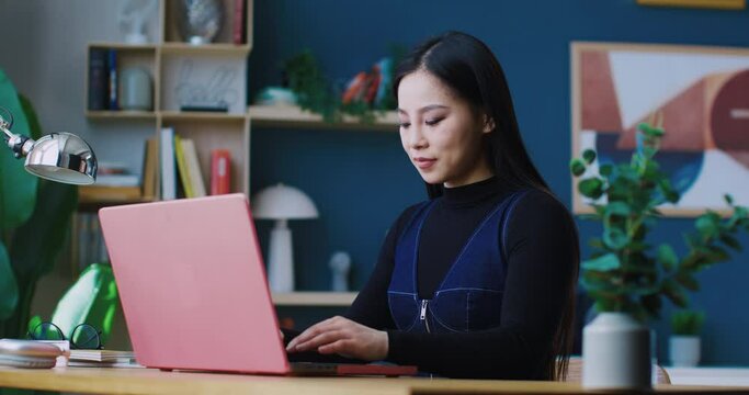 Beautiful young Asian woman with long black hair opening pink laptop. Preparing to work or study remotely. Sitting in cosy modern apartment. Entering text or information on keyboard.